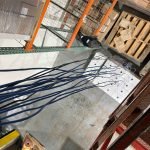 Verkada Installers Multiple blue cables are hanging vertically in a warehouse, viewed from above. Nearby are pallets, boxes, shelves, and metal racks on a concrete floor.