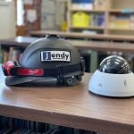 Verkada Installers A black hard hat with a headlamp and Jendy Solution Sdn Bhd label is placed on a wooden table next to a white dome-shaped security camera, with blurred bookshelves in the background.