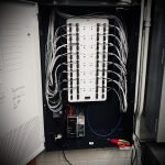 Verkada Installers A neatly organized electrical panel with multiple labeled white wires connected to circuit breakers, a power supply unit at the bottom, and a pair of green and pink-handled scissors resting inside the panel.